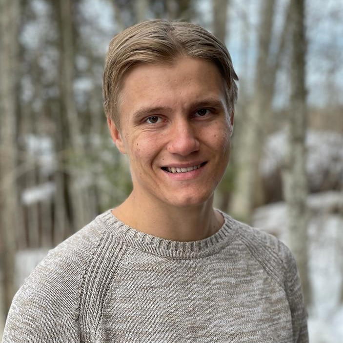 Elmeri - Portrait of a fair-haired man smiling in front of trees. He's wearing a gray sweater.