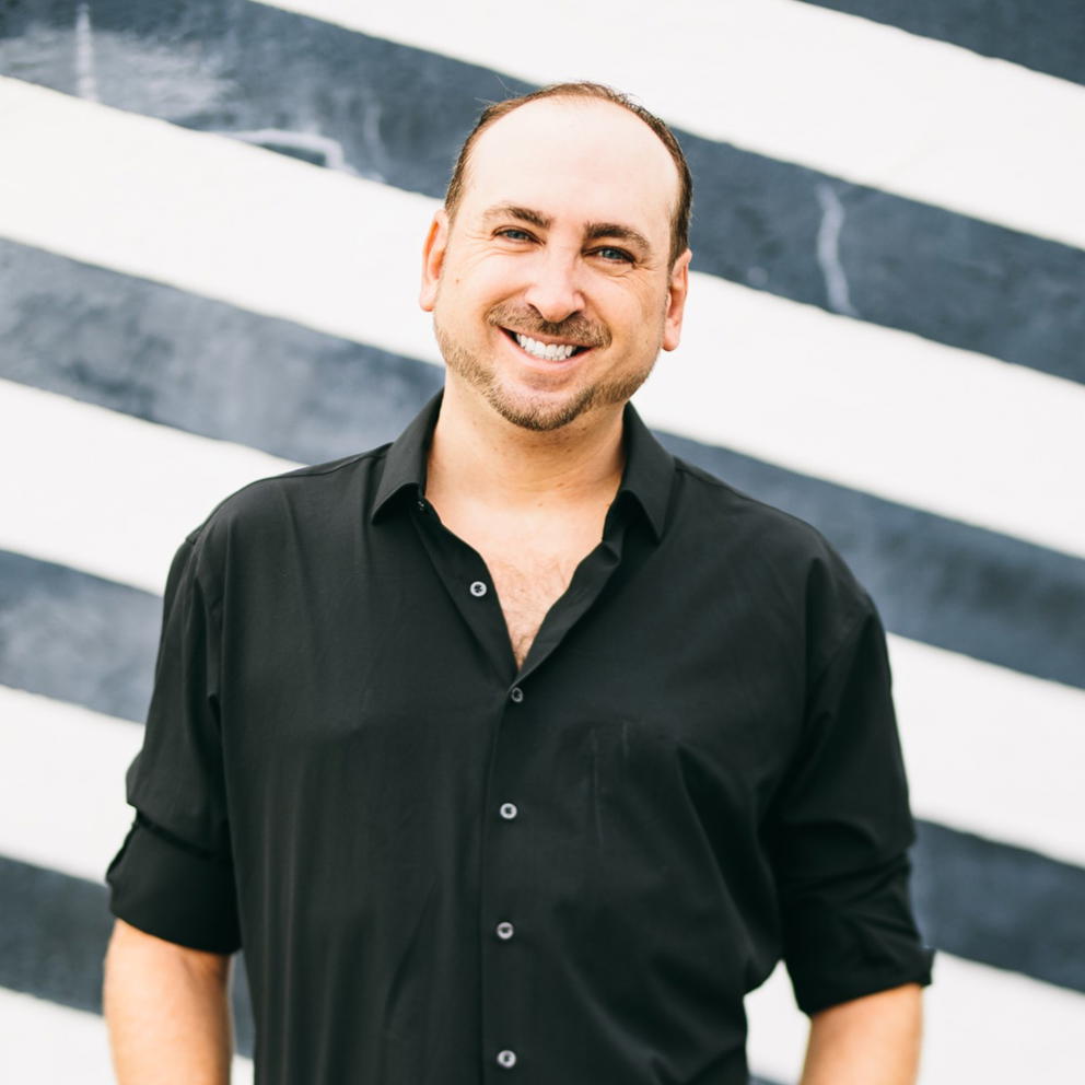 Justin Belmont - A smiling man in a black shirt stands in front of a black and white striped wall.