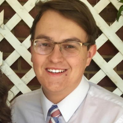 Zak Jacoby - Smiling man in glasses and tie, stands in front of white lattice.